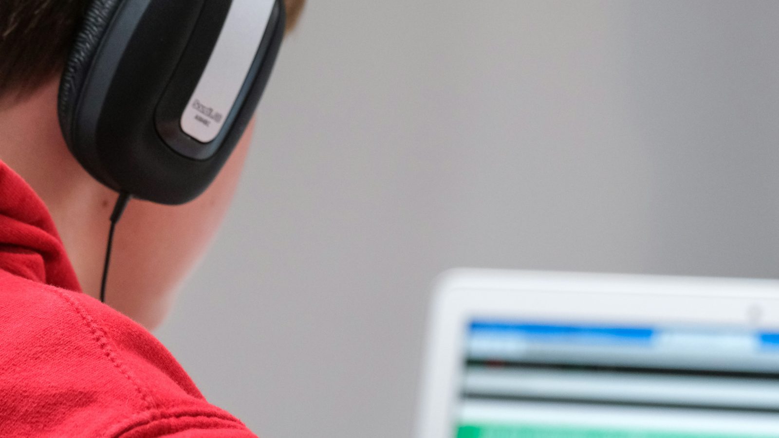 A student seen from behind wearing large over-ear headphones and a red top, holding a pen in front of an open laptop