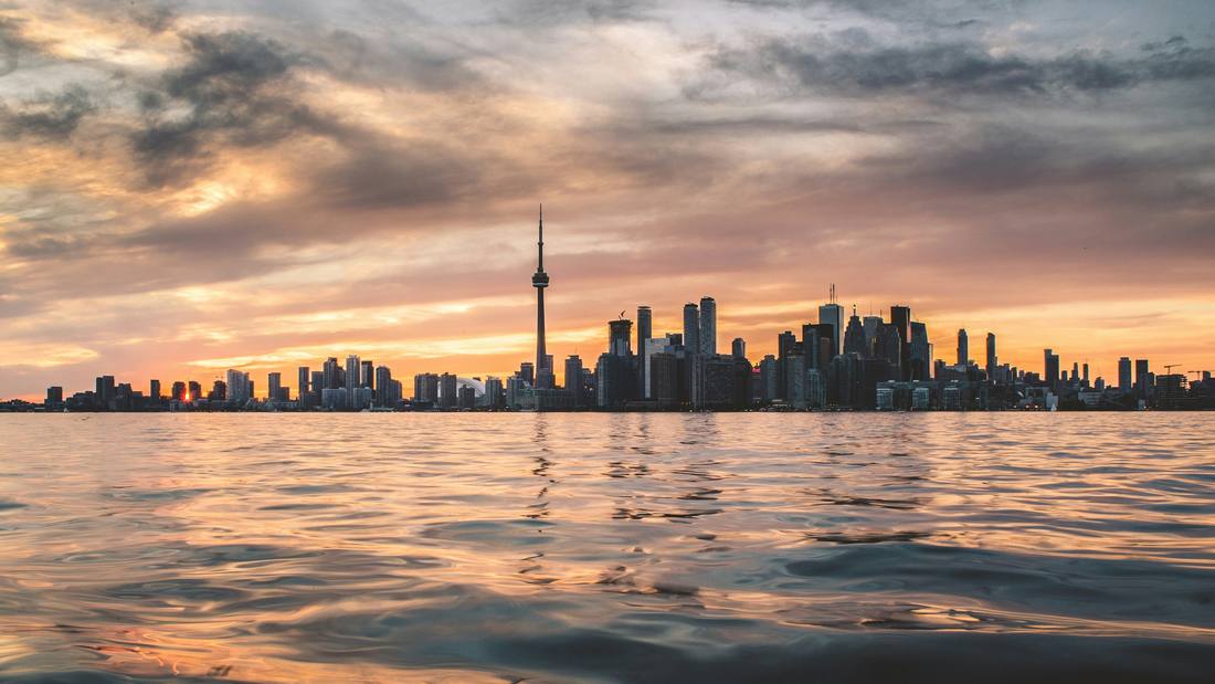 The Toronto skyline at sunset seen across the water, with the CN Tower rising above the city and golden light on the clouds