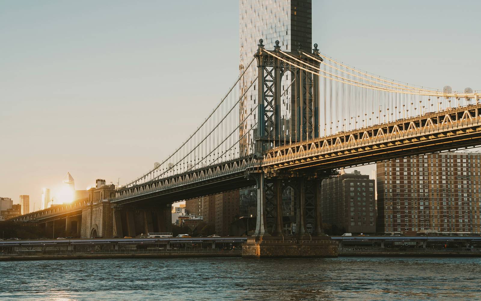 The Manhattan Bridge crossing the East River in New York at sunset, with a glass tower and brick waterfront buildings behind it