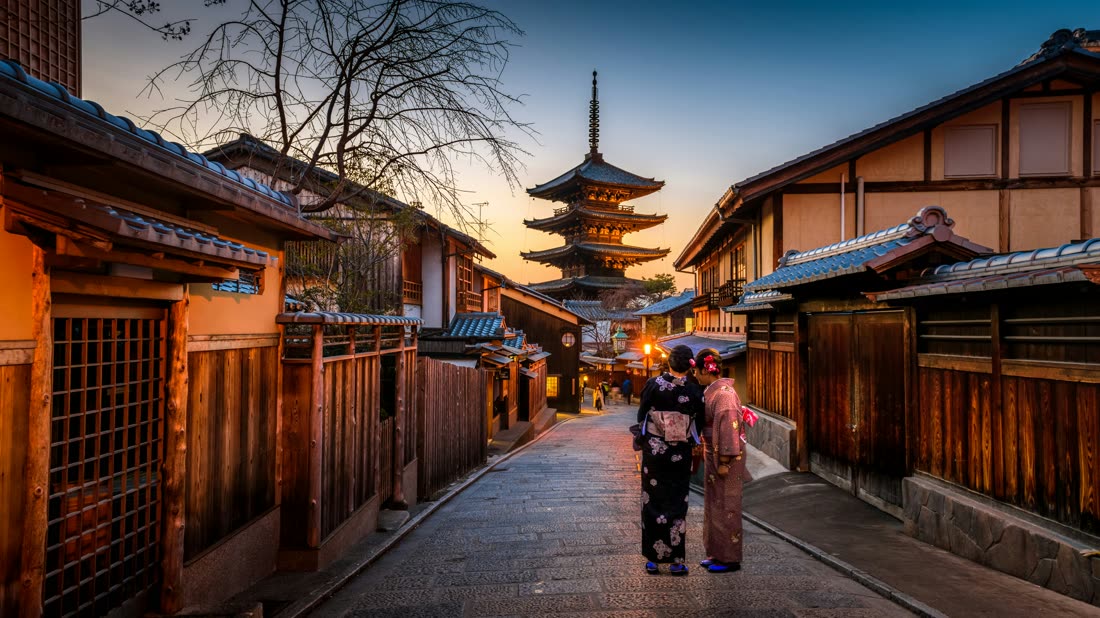 A traditional stone-paved street in Tokyo at sunset, with a tall multi-tiered pagoda at the far end and wooden townhouses lit by warm lamps on either side