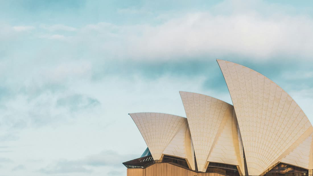 The white sail-shaped roofs of the Sydney Opera House seen close up over calm water, against a soft pale sky