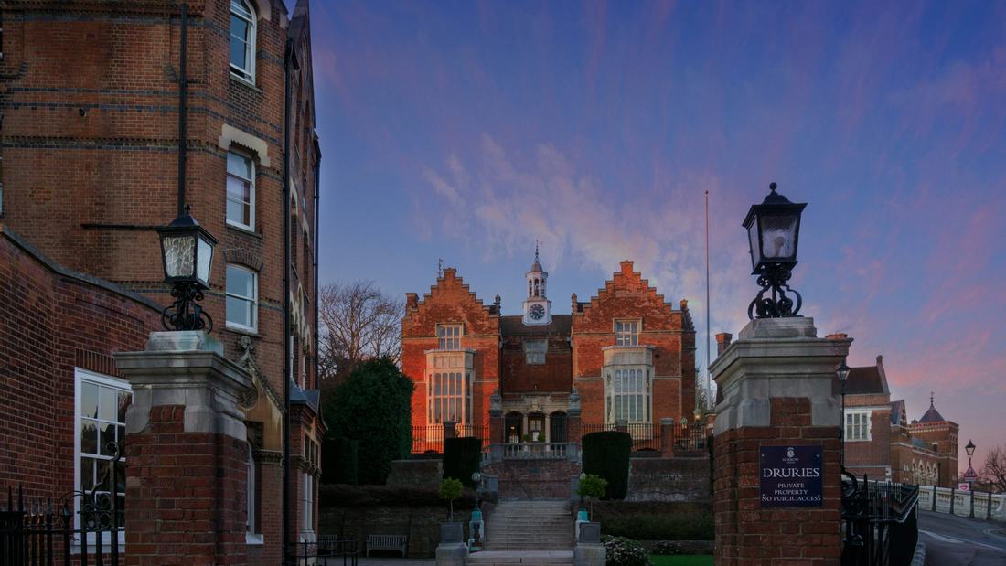 The Druries building at Harrow School at dusk, with its red-brick facade, white clock cupola and weathervane set against pink sunset clouds, framed by brick gate pillars topped with lanterns